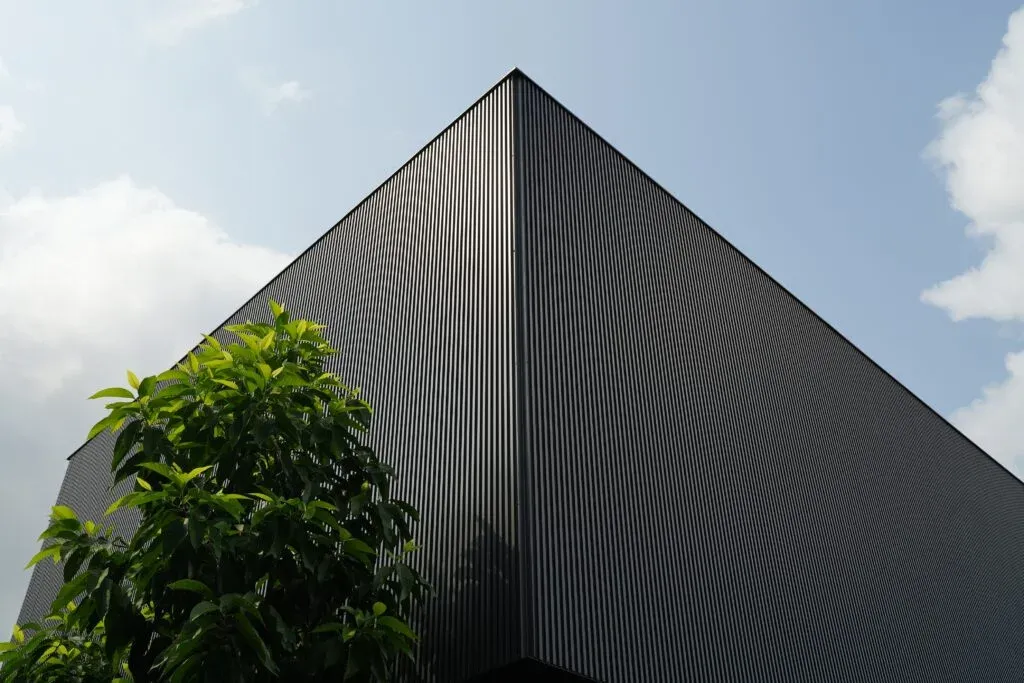 Corner of a modern black corrugated building with a leafy green tree against a blue sky.