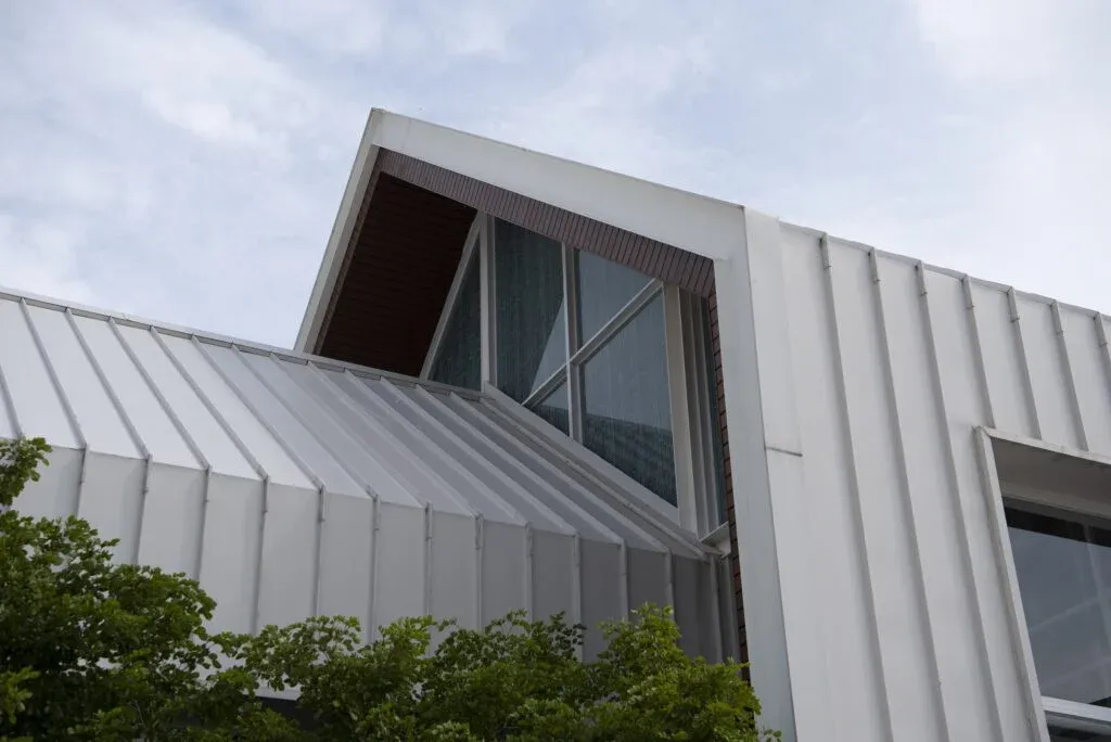 Modern building with angular rooflines and large windows, featuring metallic panels. Green foliage is visible in the foreground.