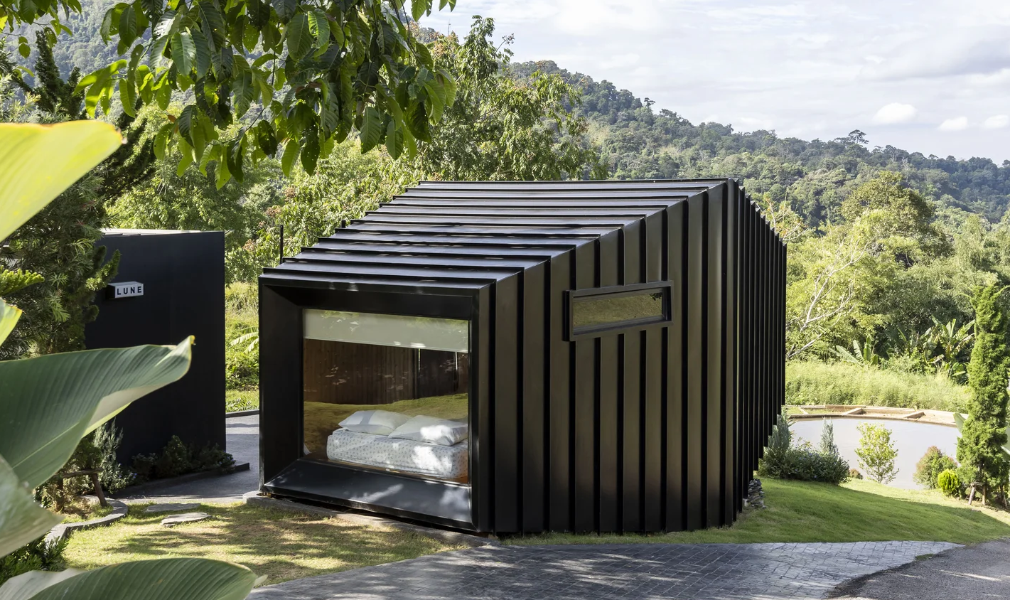 Black modern cabin with large window, set in a lush green landscape with hills in the background, under a partly cloudy sky.