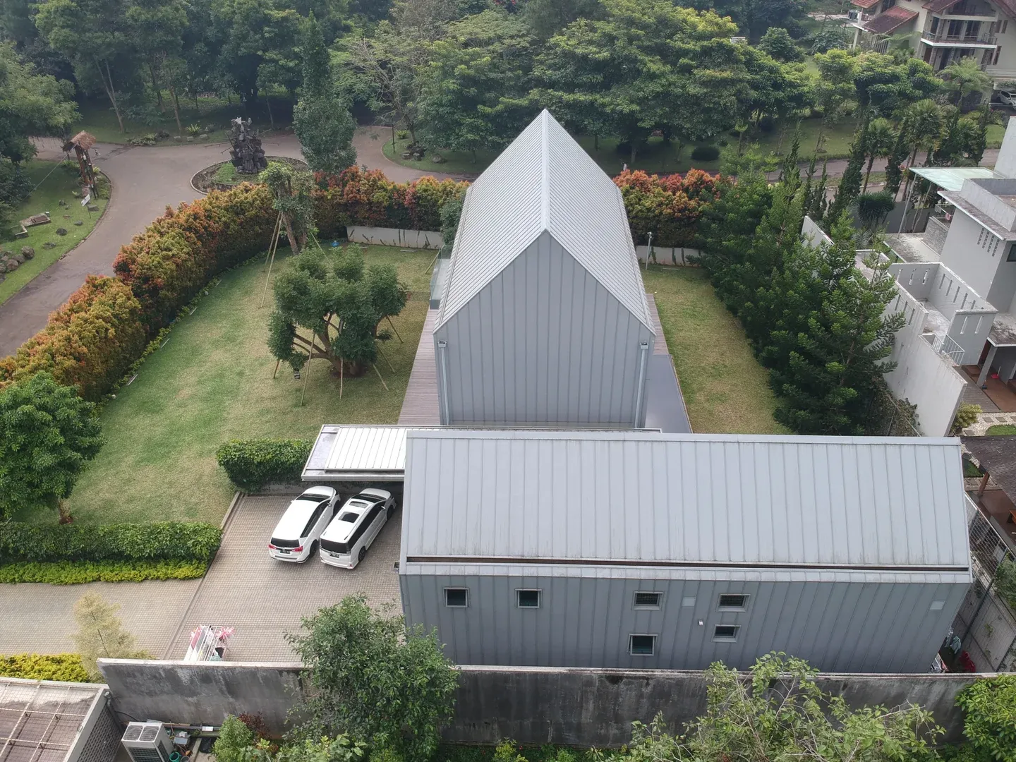 Aerial view of a modern, metal-roofed house surrounded by trees and greenery, with two cars parked in the driveway.