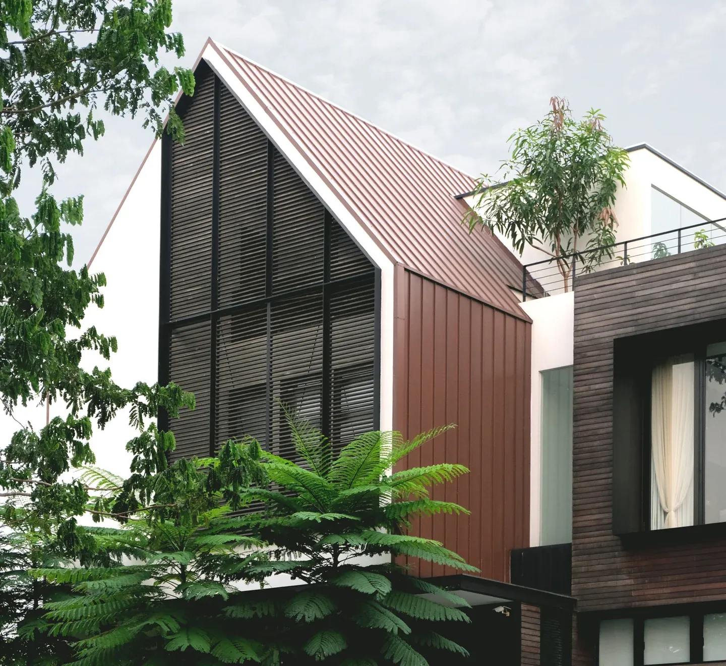 Modern house with a tall, sloped brown roof, large windows, and greenery around. The exterior combines metal and wood elements.
