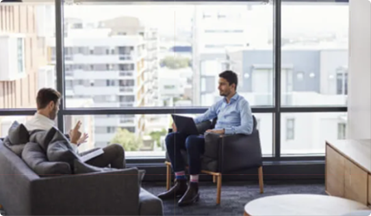 Two people having a discussion in a modern office with large windows and cityscape view. One is seated on a sofa, the other on a chair.